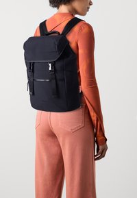 Zaino nero con top pieghevole, spallacci regolabili, tasca anteriore con zip e hardware argentato, indossato da una persona con una camicia a fantasia.