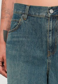 Džínsy v stredne modrej farbe, s oranžovým prešívaním, strieborným gombíkom a dvoma prednými vreckami. Tkanina sa zdá byť mierne textúrovaná.