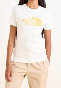 Camiseta de algodón blanca con mangas cortas que presenta un logo gráfico amarillo "THE NORTH FACE" en la parte frontal. Corte casual y textura suave.