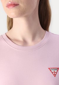 T-shirt rosa chiaro con scollo rotondo. Presenta un piccolo logo triangolare rosso e bianco sulla parte sinistra del petto, texture liscia.