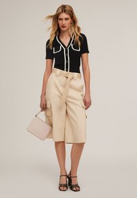 Czarna prążkowana bluzka z białym wykończeniem i złotymi guzikami, beżowe szorty cargo z wysokim stanem i paskiem oraz różowa torebka trzymana w jednej ręce.