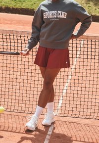 Felpa grigia con testo "SPORTS CLUB", gonna bordeaux, scarpe da ginnastica bianche e calze bianche, in piedi su un campo da tennis vicino a una rete.