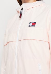 Jasnoróżowa kurtka przeciwwiatrowa z zamkiem błyskawicznym, kapturem, elastycznymi mankietami oraz naszywką z logo na przodzie. Charakteryzuje się gładką teksturą i lekkim materiałem.