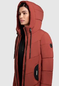 Kurtka pikowana w kolorze rdzawej czerwieni, z kapturem, zamkiem błyskawicznym i czarnymi akcentami. Teksturowana tkanina z widocznym przeszyciem i detalami kieszonek.