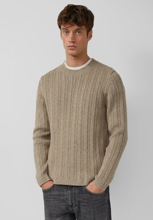 Jonge man met lichtbruin krullend haar, gekleed in een beige ribbelgebreide trui en donkere spijkerbroek, met zijn handen in zijn zakken, neutrale grijze achtergrond.
