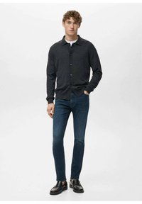 Cardigan gris oscuro abotonado con cuello, combinado con jeans oscuros ajustados y zapatos negros sin cordones, llevado por una figura masculina.