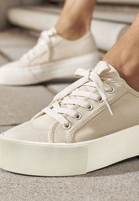 Beige kangastossud ümmarguse varbaga, lamedate paeltega disain ja tekstureeritud kummitald. Omab metallist silmuseid ja õmmeldud detaile.