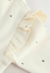 Cremefarvet jumpsuit med sort hjerte mønster, der har flæseærmedetalje og trykknaplukning for nem påklædning. Blød strikstruktur.