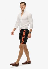 Pantaloni da nuoto neri con vivace branding arancione "SUPERDRY", cintura elasticizzata con cordino, abbinati a una camicia di lino bianca e mocassini marroni.