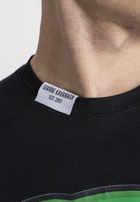 Černé tričko s úzkým střihem, na límci má tkanou nálepku s nápisem "Gianni Kavanagh EST. 2017". Hladká textura, bez vzorů.