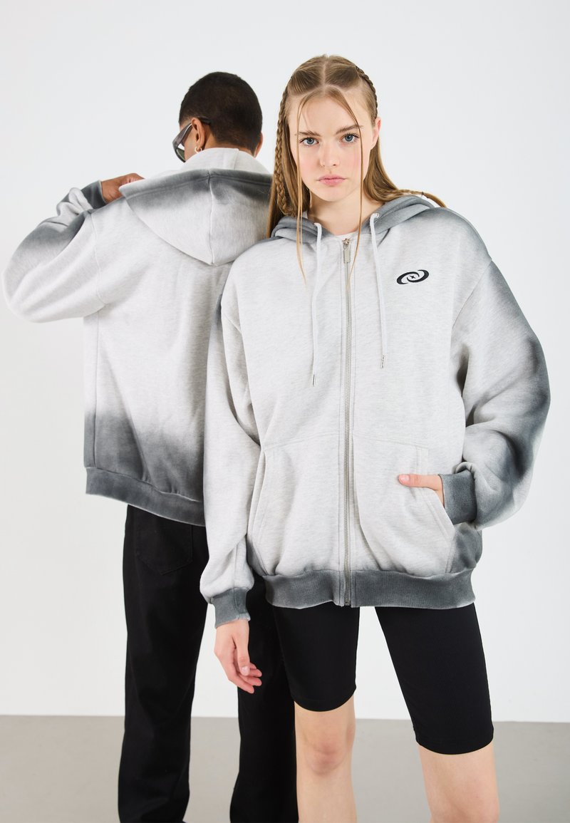 Felpa grigia con zip e un gradiente ombre da grigio chiaro a grigio scuro. Presenta un logo sul petto sinistro e tasche frontali. Il modello indossa pantaloni corti neri.