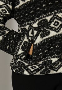 Mano in una giacca in pile a fantasia bianco e nero con tasca visibile con zip nera e un design geometrico sul tessuto della giacca.