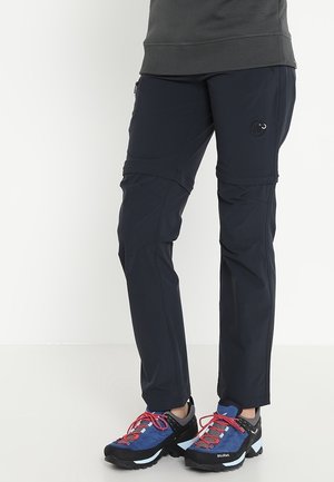 Person, die marineblaue wandelbare Wanderhose und blau-schwarze Trailschuhe mit roten Schnürsenkeln trägt, steht auf einem weißen Boden.