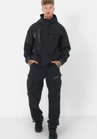 Giacca e pantaloni impermeabili neri con molteplici tasche, cappuccio regolabile e una texture liscia. Presenta il logo sui pantaloni e cerniere.