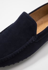Marineblå ruskinds slip-on sko med rund tå, stikninger og en glat inderside. Tekstureret sål med små gummidutter.