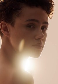 Close-up van een persoon met kort, krullend haar en een zichtbare oorbel, verlicht door een zachte, gloedvolle licht van achteren.