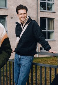 Sort hættetrøje med beige for, båret over et sort basislag, parret med lyseblå jeans. En lille crossover-taske er synlig.