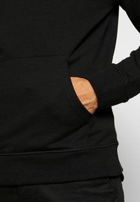 Czarna bluza z kapturem z kieszenią kangurka, ściągaczami przy mankietach i u dołu. Gładka tekstura z detalami szwów, pokazana z ręką w kieszeni.