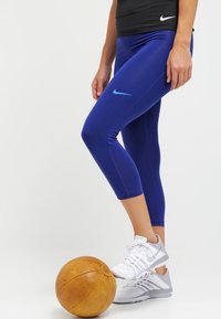 Námořnické modré capri legíny se světle modrým logem Nike, vyrobené z hladkého, elastického materiálu. Nosené s bílými sportovními botami vedle hnědého basketbalového míče.