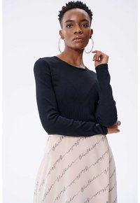 Schwarzes langärmliges Rippoberteil mit rundem Ausschnitt, kombiniert mit einem beigen Rock, der ein wiederholtes schwarzes Schriftmuster aufweist.