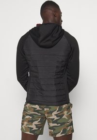Black hooded jacket with padded back, featuring a smooth texture and fitted design. Worn with camo shorts.
