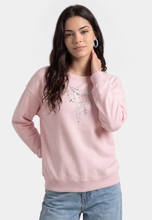Giovane donna con lunghi capelli scuri che indossa una felpa rosa chiaro con motivo a catena argento e orecchini a cerchio blu, posando con la mano sul collo.