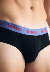 Čierne trenky so svetlomodrým pásom, na ktorom je nápis "HUGO" v červenej farbe. Tkanina vyzerá hladko a elasticky, navrhnutá pre pohodlie a dobré padnutie.