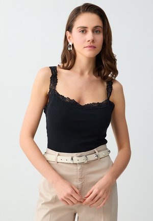 Kobieta o brązowych włosach do ramion, nosząca srebrne kolczyki koła, czarny top wykończony koronką, beżowe spodnie z wysokim stanem oraz biały pasek.