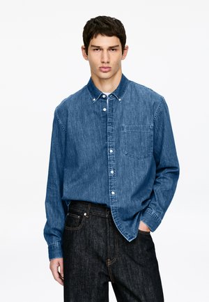 Junger Mann mit kurzen dunklen Haaren, der ein blaues Jeanshemd mit Knöpfen und schwarze Jeans trägt und mit einer Hand in der Tasche steht.