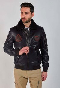 Homme avec une barbe portant une veste bombardier en cuir noir avec des écussons et un pantalon cargo beige, ajustant sa fermeture éclair sur un fond uni.