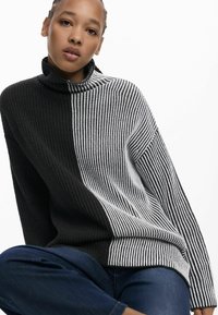 Ce pull en maille épaisse présente un col montant, des motifs rayés alternant noir et blanc, et une coupe ample. Texturé et chaud, il est idéal pour superposer.