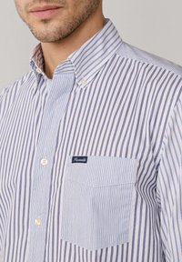 Camicia con bottoni in bianco e blu a righe verticali. Presenta un colletto, una tasca frontale e un'etichetta del marchio. Materiale in cotone con una consistenza liscia.