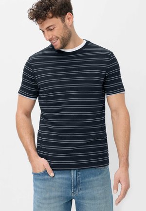 Uomo con capelli corti e ricci che indossa una maglietta a righe bianche e nere e jeans blu, sorride e guarda verso il basso, una mano in tasca.