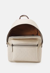 Mały beżowy skórzany plecak z tłoczonym wzorem, przednia kieszeń na suwak, otwarta główna przegroda pokazująca brązowe materiałowe wnętrze oraz wewnętrzną kieszeń na suwak.