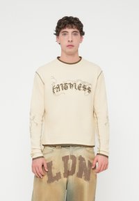 Κρεμ πουκάμισο μακρυμάνικο με εφαρμοστό καφέ γιακά, με γραφικό κείμενο "Faintless" και διακοσμητικά σχέδια στα μανίκια, φορεμένο με ξεθωριασμένο τζιν παντελόνι με το γράμμα "LDN".