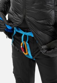 Personne portant une veste noire et un pantalon, en train de fixer un harnais d'escalade bleu avec un mousqueton orange autour de la taille.