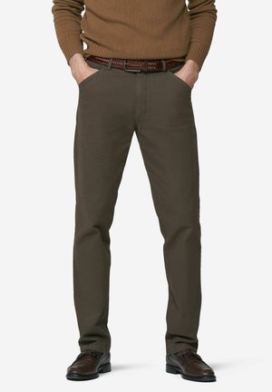 Mann trägt braunen Strickpullover, olivgrüne gerade geschnittene Hose mit braunem geflochtenem Gürtel und dunkelbraune Lederschuhe, Hände in den Taschen.