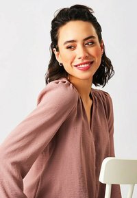 Blusa in morbido tessuto malva con maniche lunghe, scollatura a V e delicate pieghe sulle spalle; il modello è seduto accanto a una sedia bianca.