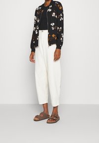 Chaqueta bomber negra con bordado floral, combinada con pantalones anchos blancos y sandalias marrones. Los tejidos son suaves; el diseño presenta un cierre de cremallera.