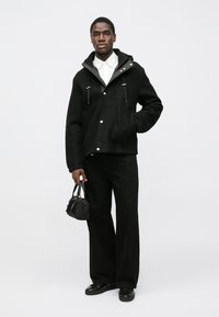 Sort fleecejakke med hætte, frontlommer med klap og snoredetaljer, kombineret med sorte bukser med vide ben og en lille håndtaske.