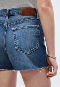 Κλείσιμο πλάνου ενός ατόμου που φοράει μπλε τζιν σορτς Calvin Klein με πίσω τσέπη και δερμάτινη ταμπέλα με το λογότυπο, καθώς και ασημένιο βραχιόλι στον καρπό.