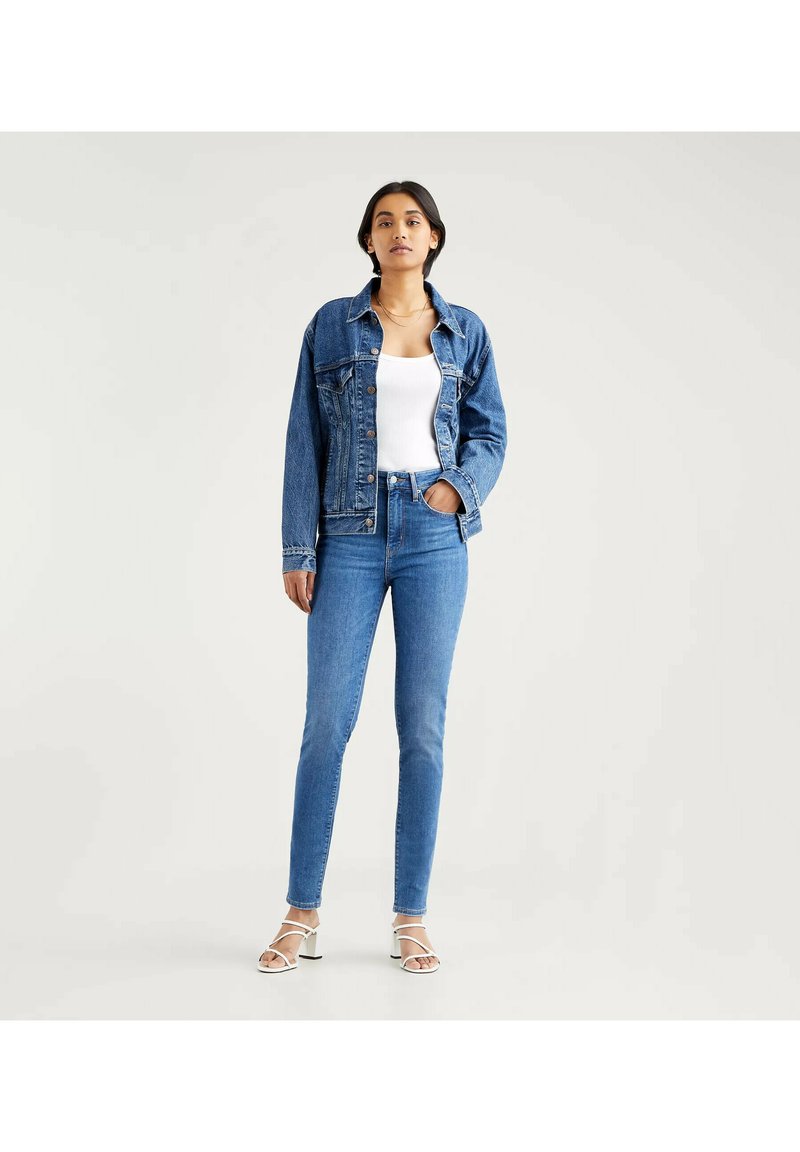 Blaue Jeansjacke über einem weißen ärmellosen Oberteil, kombiniert mit hochtaillierten blauen Jeans. Das Modell trägt weiße Sandalen mit Riemen.