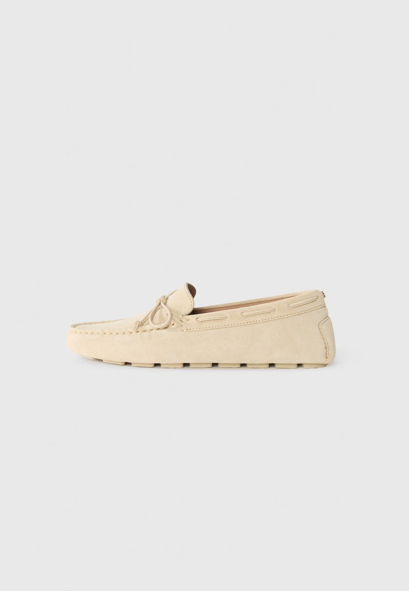 Mocassin en suède beige avec un bout rond, des détails de lacets à l'avant et une semelle texturée avec des découpes rectangulaires.