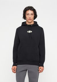Giovane con capelli scuri di lunghezza media che indossa un hoodie nero con un piccolo logo ovale e pantaloni felpa grigi con una toppa sulla coscia sinistra.
