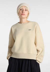 Sudadera beige con un corte relajado, puños acanalados y un pequeño logo en el pecho. Llevada con un gorro de punto de color claro.