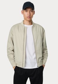 Lichtbeige bomberjack met een geribbelde kraag en manchetten, gladde textuur, vooraan een rits, gedragen over een wit T-shirt, gecombineerd met zwarte broek.