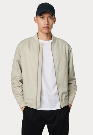 Giacca bomber beige chiaro con colletto e polsini a costine, texture liscia, zip frontale, indossata sopra una maglietta bianca, abbinata a pantaloni neri.