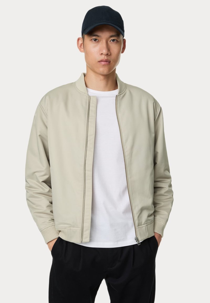 Lichtbeige bomberjack met een geribbelde kraag en manchetten, gladde textuur, vooraan een rits, gedragen over een wit T-shirt, gecombineerd met zwarte broek.