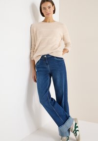 Suéter beige de punto acanalado con rayas blancas, combinado con vaqueros azul de pierna ancha, puños remangados y zapatillas con acentos verdes.