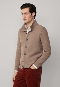 Bežový pletený cardigan s vysokým golierom, ktorý má tmavé gombíky, štruktúrovanú tkaninu a rebrované manžety. V kombinácii s červenými kordových nohavicami.